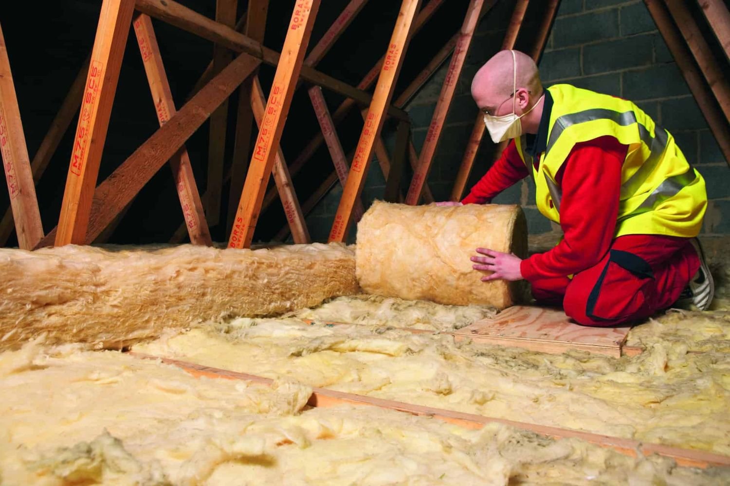 A man in a yellow vest and safety gear installs insulation in an attic, ensuring proper energy efficiency and safety.