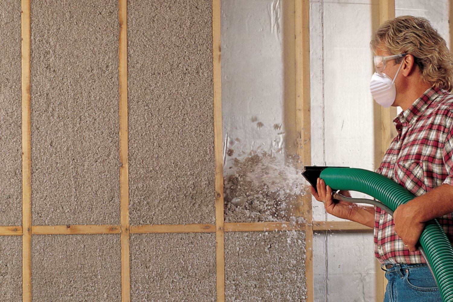 A person is using a green hose to spray insulation material into wall framing, demonstrating a home renovation process.