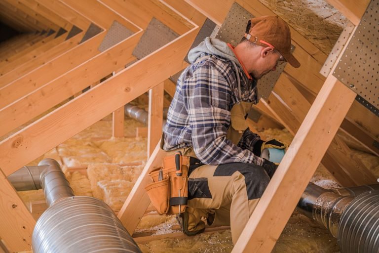 A worker in a plaid shirt and tool belt crouches in an attic, inspecting ducts and insulation amid wooden beams.