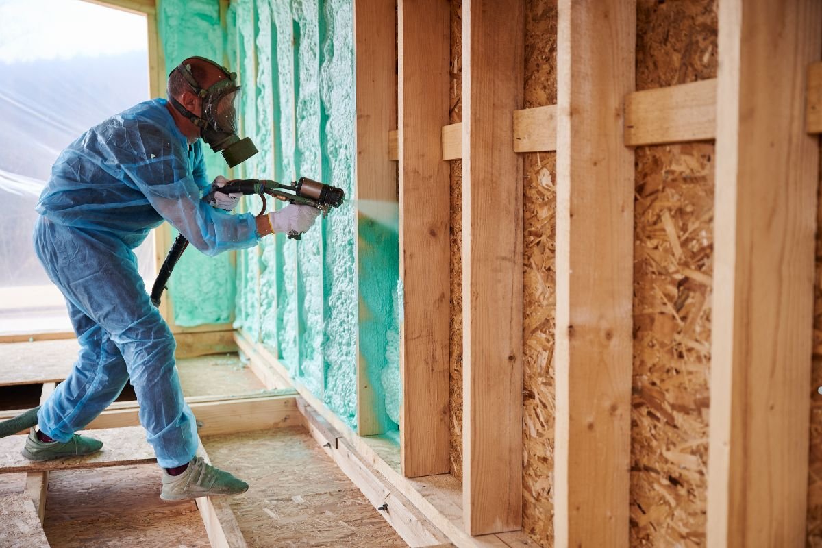 A worker, dressed in protective gear, sprays insulation foam on wooden beams inside a construction site.