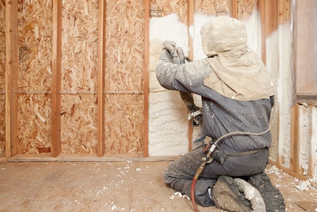 A worker applies foam insulation to wall framing, wearing protective gear and kneeling on the floor in a construction setting.