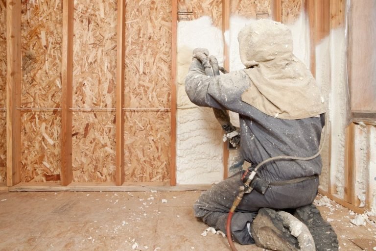 A worker applies foam insulation to wall framing, wearing protective gear and kneeling on the floor in a construction setting.